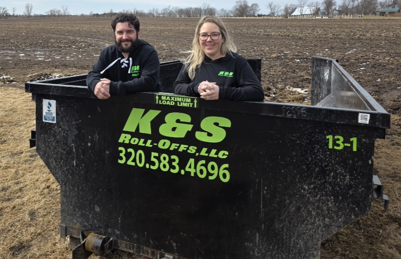 two people standing in a k&s roll-off dumpster in a field